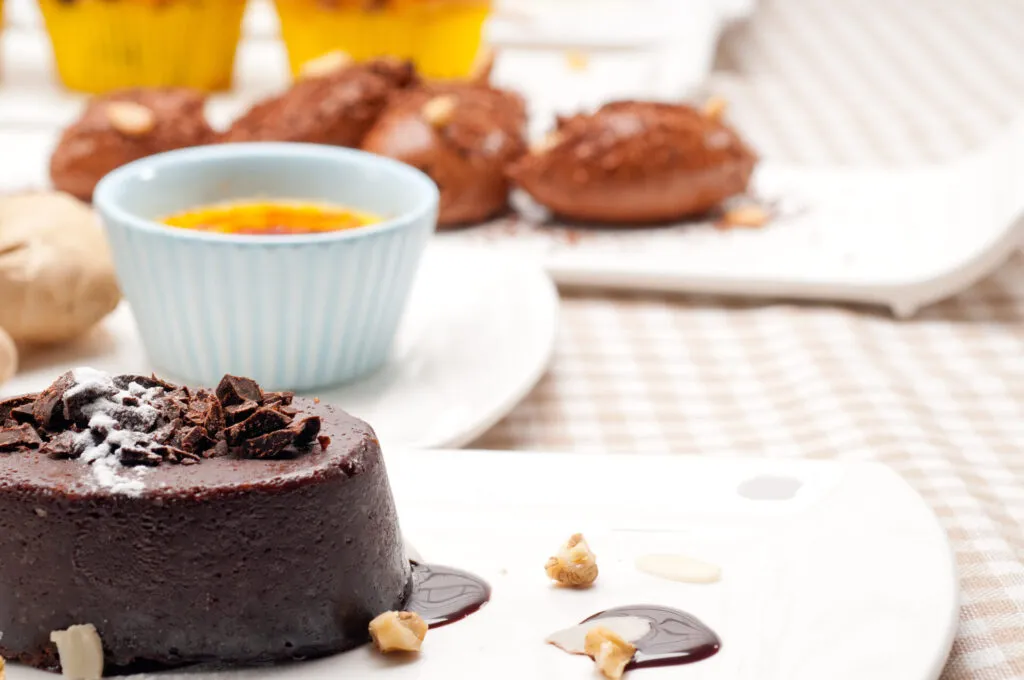 Bonet piemontese al cioccolato servito su un piatto bianco, decorato con scaglie di cioccolato, salsa al cacao e granella di nocciole.