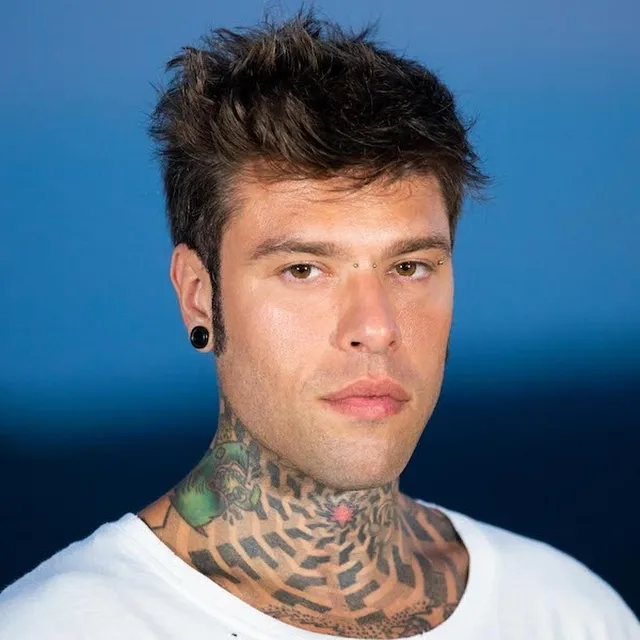Ritratto di Fedez con maglietta bianca, capelli scuri spettinati e tatuaggi visibili sul collo, davanti a uno sfondo blu sfumato.