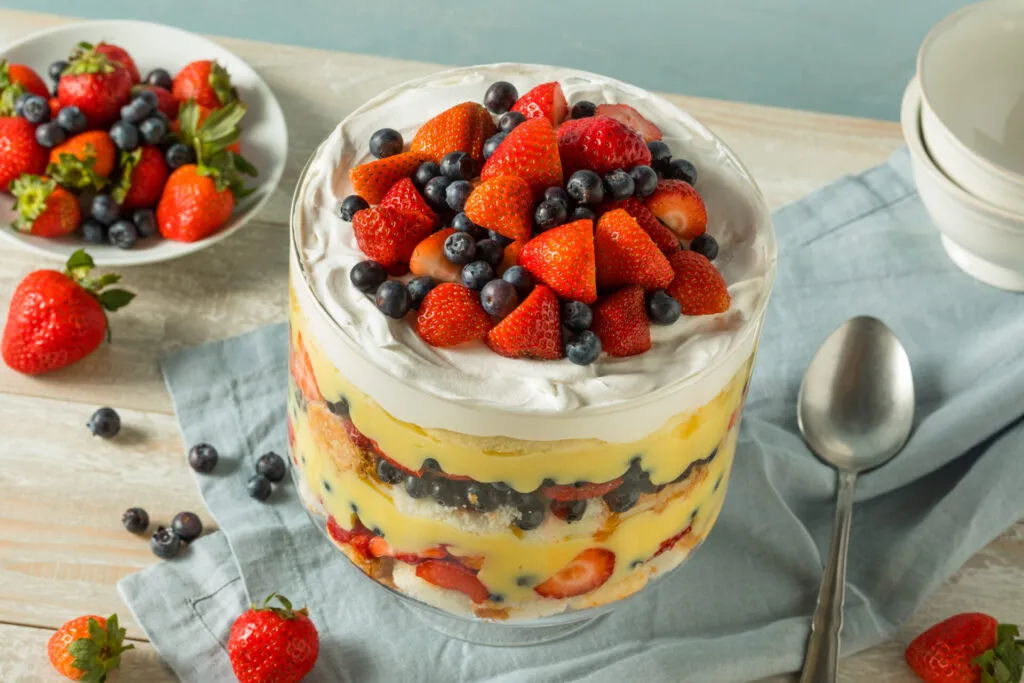 Zuppa inglese in coppa di vetro con strati di crema pasticcera, pan di Spagna, fragole, mirtilli e panna montata, decorata con frutta fresca.