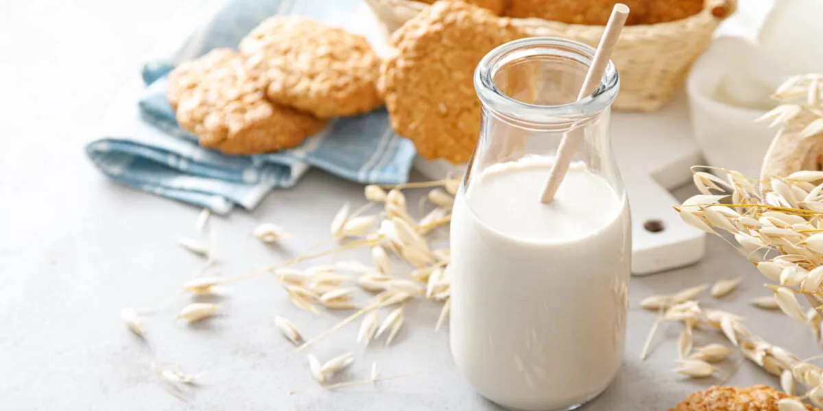 Bottiglia di bevanda d’avena con cannuccia, circondata da spighe, fiocchi d’avena, biscotti e pane integrale.