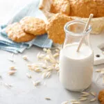 Bottiglia di bevanda d’avena con cannuccia, circondata da spighe, fiocchi d’avena, biscotti e pane integrale.
