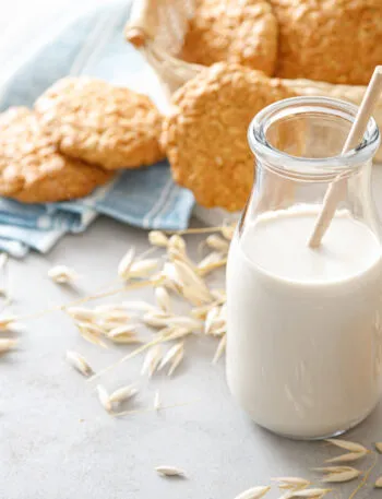 Bottiglia di bevanda d’avena con cannuccia, circondata da spighe, fiocchi d’avena, biscotti e pane integrale.