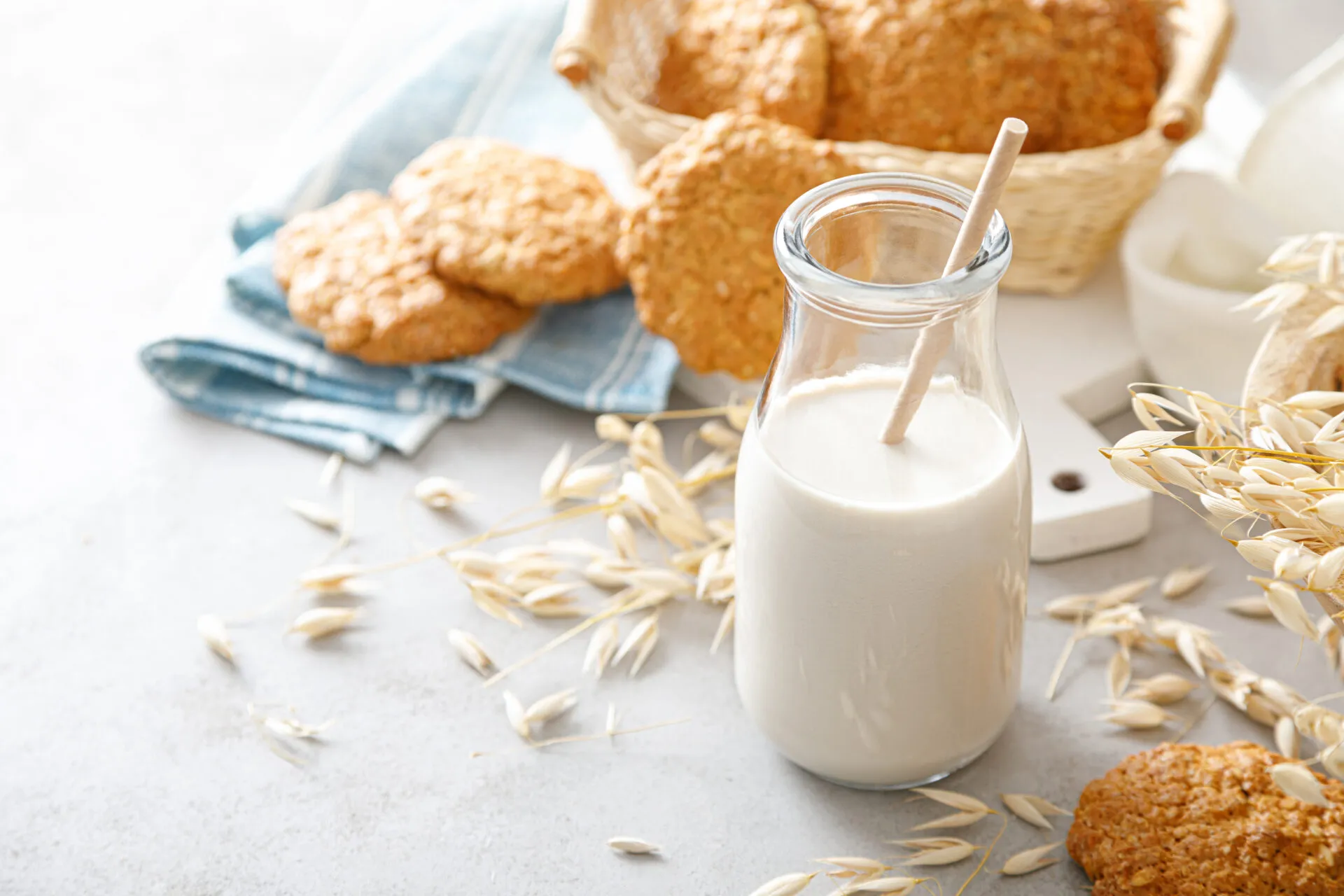 Bottiglia di bevanda d’avena con cannuccia, circondata da spighe, fiocchi d’avena, biscotti e pane integrale.