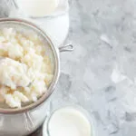 Grani di kefir di latte in un colino con cucchiaio, accanto a barattoli di latte su fondo chiaro.
