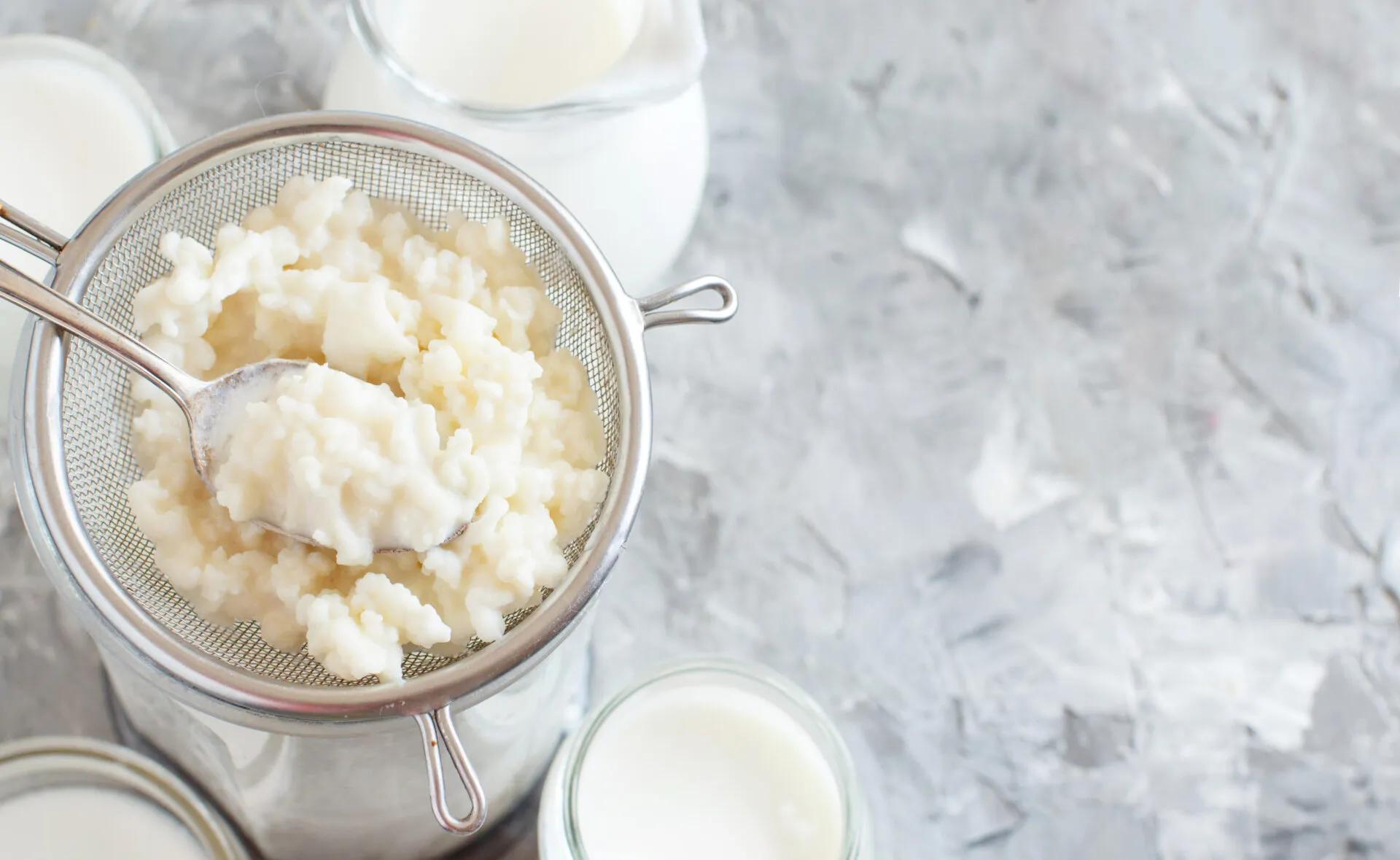 Grani di kefir di latte in un colino con cucchiaio, accanto a barattoli di latte su fondo chiaro.