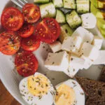Piatto per colazione salata fit con uova sode, avocado, pomodorini, cetrioli, formaggio a cubetti e pane integrale.