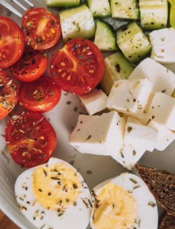 Piatto per colazione salata fit con uova sode, avocado, pomodorini, cetrioli, formaggio a cubetti e pane integrale.