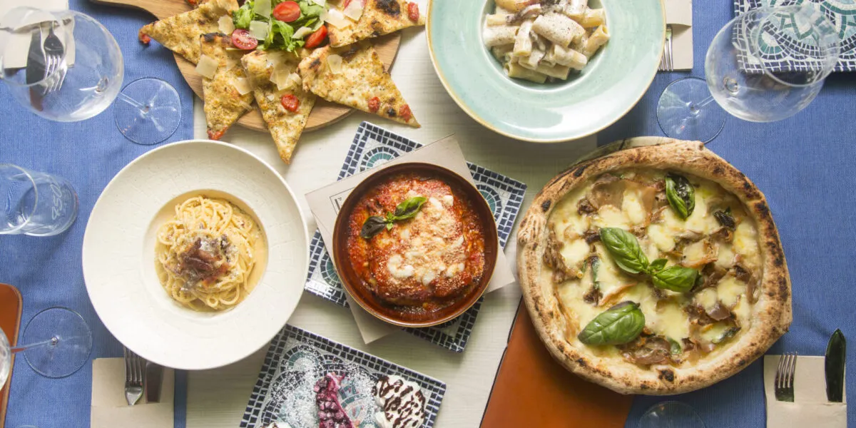 Tavola con piatti della cucina regionale italiana, tra pizza, pasta, parmigiana, focaccia e dolci.