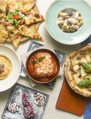 Tavola con piatti della cucina regionale italiana, tra pizza, pasta, parmigiana, focaccia e dolci.