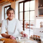 Anziana donna con grembiule prepara un impasto in cucina usando farina, uova e utensili, simbolo del cucinare senza bilancia.