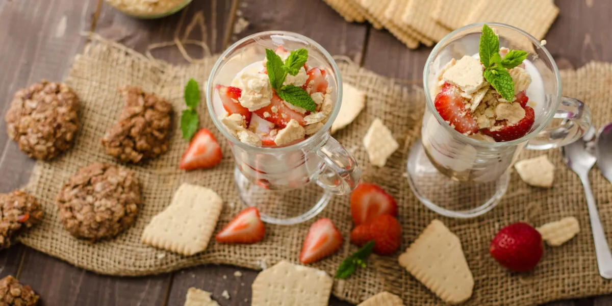 Due bicchieri di dessert proteico allo yogurt con fragole, biscotti sbriciolati e menta su tavolo in legno.