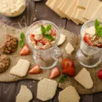 Due bicchieri di dessert proteico allo yogurt con fragole, biscotti sbriciolati e menta su tavolo in legno.