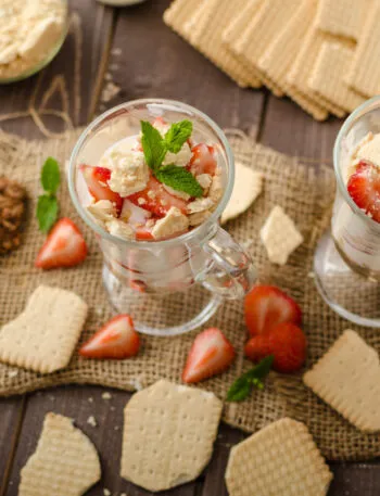 Due bicchieri di dessert proteico allo yogurt con fragole, biscotti sbriciolati e menta su tavolo in legno.