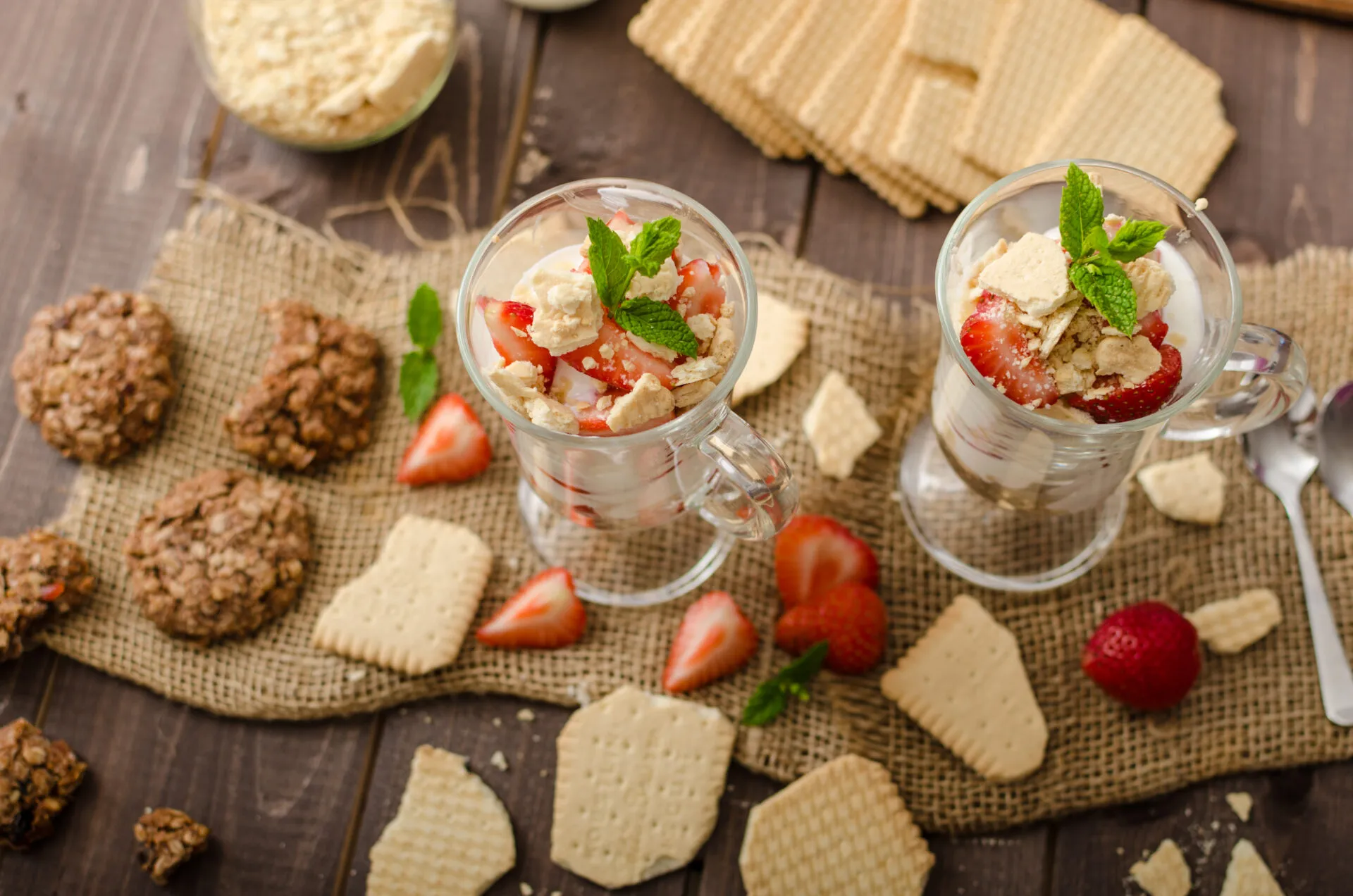 Due bicchieri di dessert proteico allo yogurt con fragole, biscotti sbriciolati e menta su tavolo in legno.