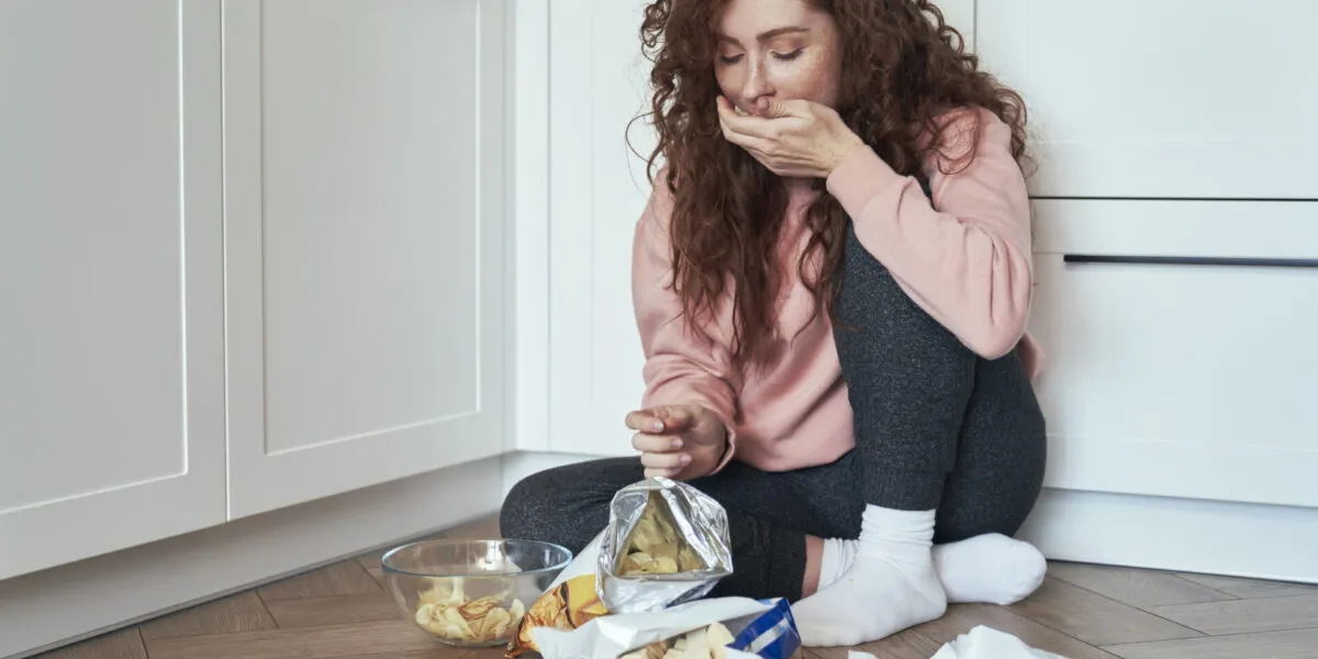 Donna seduta sul pavimento della cucina mentre mangia snack per fame nervosa, esempio di alimentazione emotiva causata da stress e ansia.