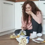Donna seduta sul pavimento della cucina mentre mangia snack per fame nervosa, esempio di alimentazione emotiva causata da stress e ansia.