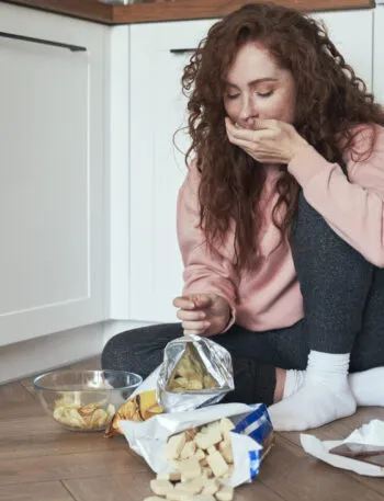 Donna seduta sul pavimento della cucina mentre mangia snack per fame nervosa, esempio di alimentazione emotiva causata da stress e ansia.