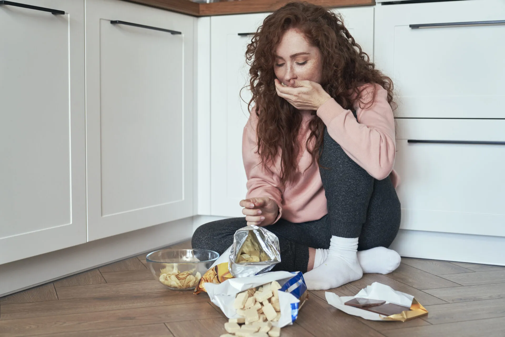 Donna seduta sul pavimento della cucina mentre mangia snack per fame nervosa, esempio di alimentazione emotiva causata da stress e ansia.