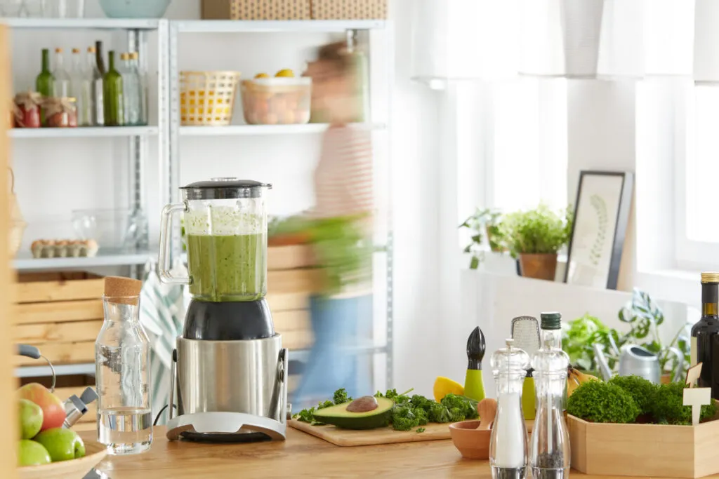 Frullatore con smoothie verde su un piano di lavoro ordinato, circondato da avocado, cavolo riccio, mele e utensili da cucina.