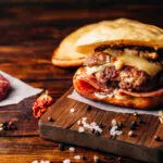 Hamburger fatto in casa con carne, formaggio fuso, funghi e prosciutto su tagliere di legno.