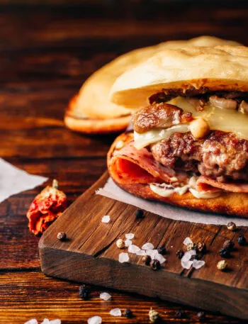 Hamburger fatto in casa con carne, formaggio fuso, funghi e prosciutto su tagliere di legno.