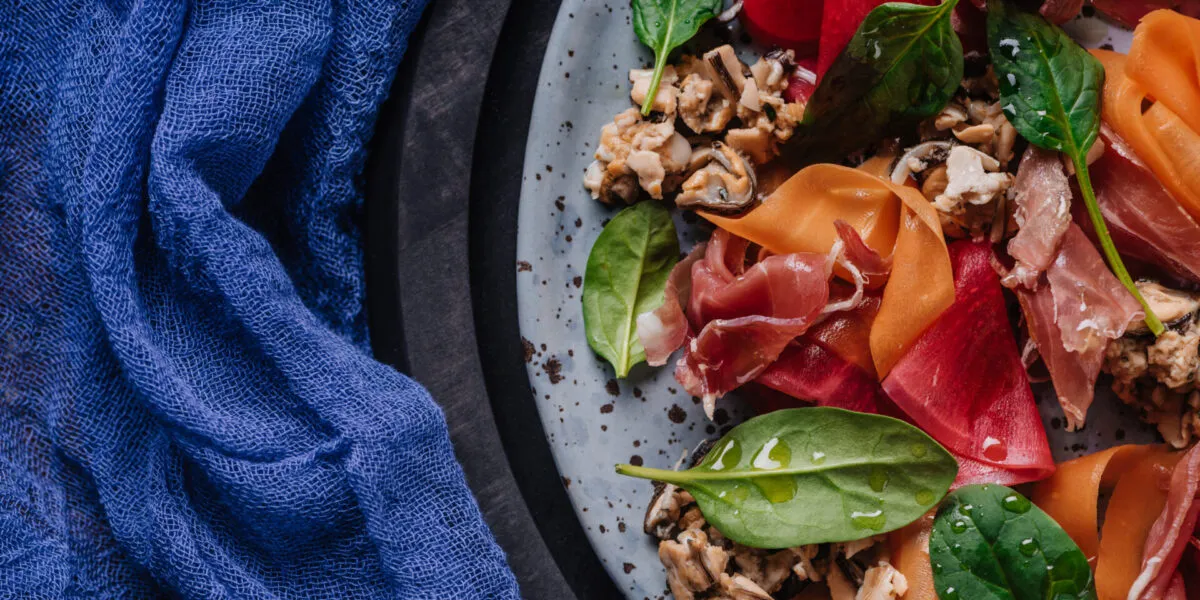 Insalata proteica con prosciutto crudo, molluschi, spinacino e verdure rosse e arancioni tagliate a nastri.