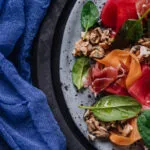 Insalata proteica con prosciutto crudo, molluschi, spinacino e verdure rosse e arancioni tagliate a nastri.