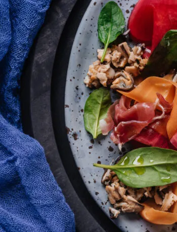 Insalata proteica con prosciutto crudo, molluschi, spinacino e verdure rosse e arancioni tagliate a nastri.
