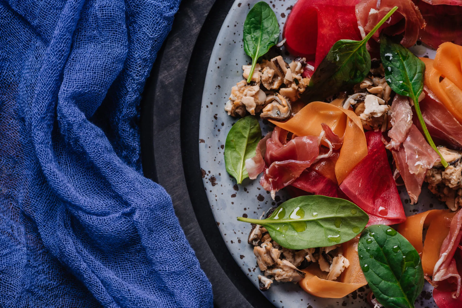 Insalata proteica con prosciutto crudo, molluschi, spinacino e verdure rosse e arancioni tagliate a nastri.