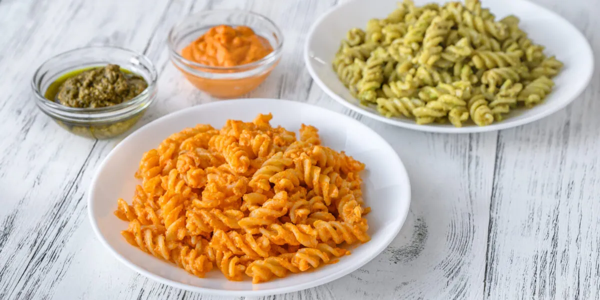 Due piatti di fusilli con condimenti cremosi, uno arancione e uno verde, accompagnati da ciotoline di pesto e salse leggere.