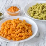 Due piatti di fusilli con condimenti cremosi, uno arancione e uno verde, accompagnati da ciotoline di pesto e salse leggere.