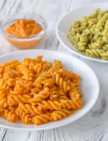 Due piatti di fusilli con condimenti cremosi, uno arancione e uno verde, accompagnati da ciotoline di pesto e salse leggere.