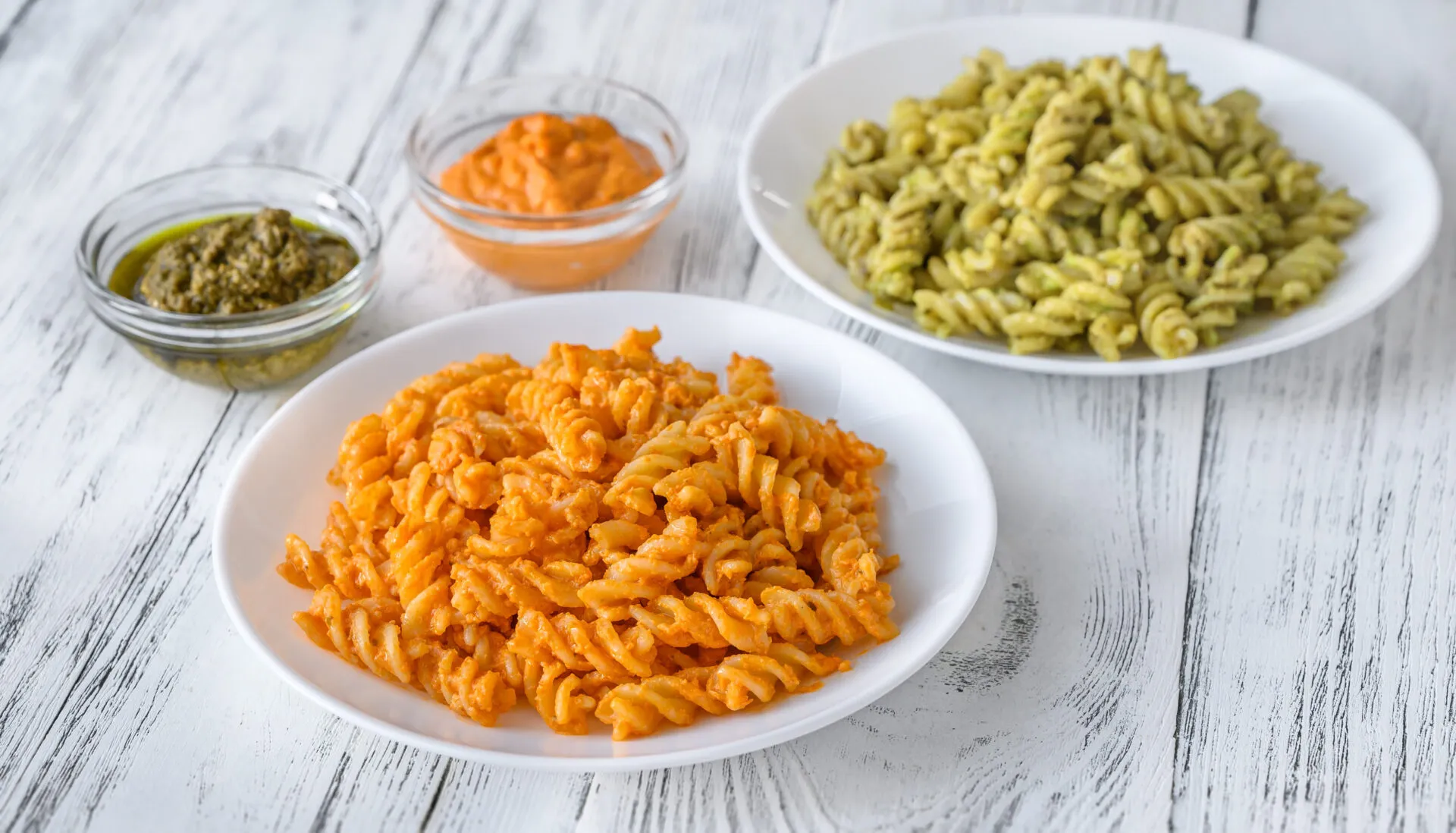 Due piatti di fusilli con condimenti cremosi, uno arancione e uno verde, accompagnati da ciotoline di pesto e salse leggere.