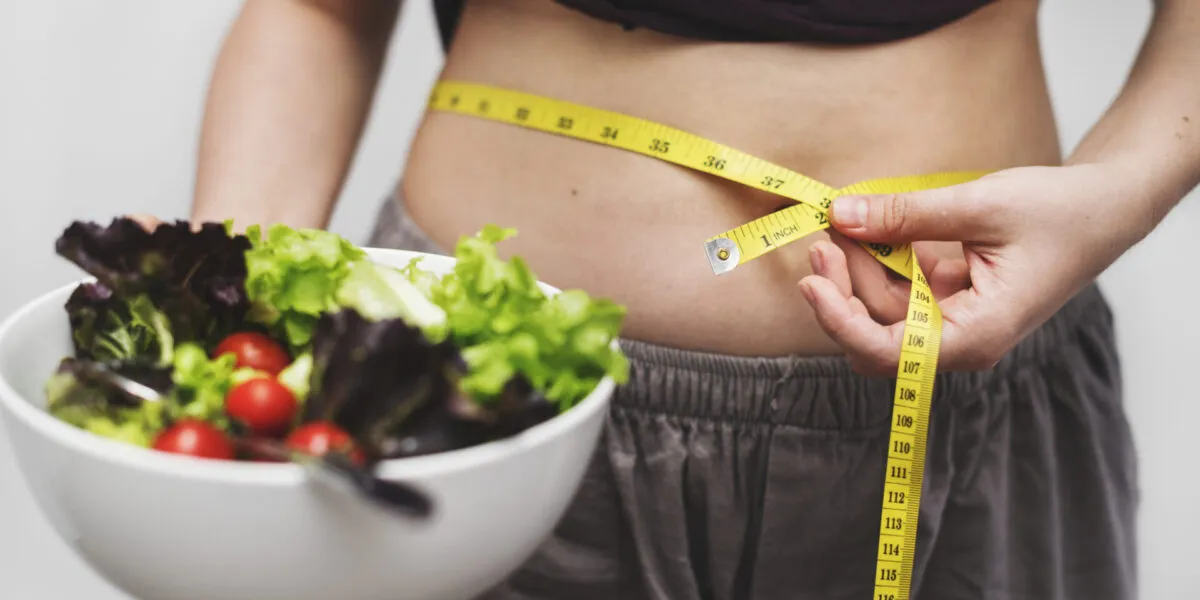 Persona che misura il girovita con un metro da sarta mentre tiene una ciotola di insalata, simbolo delle difficoltà nel seguire una dieta e costruire abitudini alimentari sostenibili.