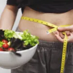 Persona che misura il girovita con un metro da sarta mentre tiene una ciotola di insalata, simbolo delle difficoltà nel seguire una dieta e costruire abitudini alimentari sostenibili.