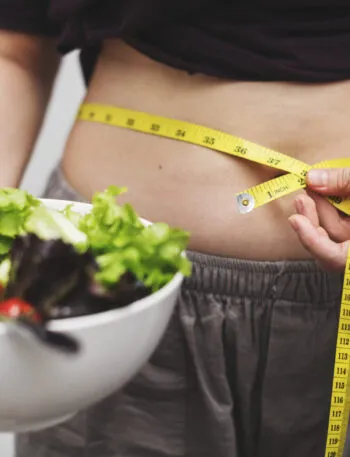 Persona che misura il girovita con un metro da sarta mentre tiene una ciotola di insalata, simbolo delle difficoltà nel seguire una dieta e costruire abitudini alimentari sostenibili.