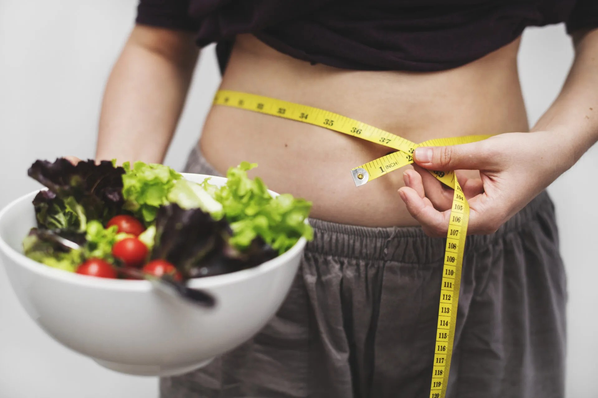 Persona che misura il girovita con un metro da sarta mentre tiene una ciotola di insalata, simbolo delle difficoltà nel seguire una dieta e costruire abitudini alimentari sostenibili.
