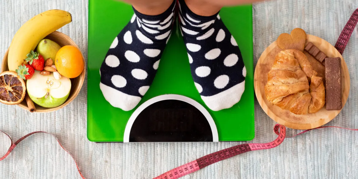 Persona sulla bilancia con a confronto alimenti sani e dolci, simbolo dello stallo del peso durante una dieta.
