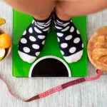 Persona sulla bilancia con a confronto alimenti sani e dolci, simbolo dello stallo del peso durante una dieta.