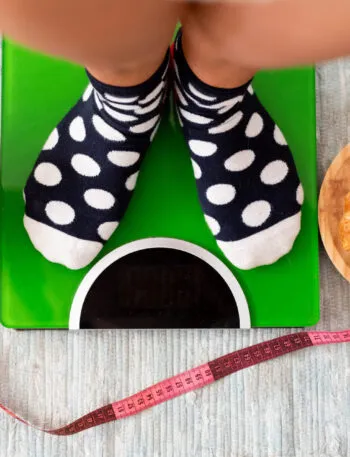 Persona sulla bilancia con a confronto alimenti sani e dolci, simbolo dello stallo del peso durante una dieta.