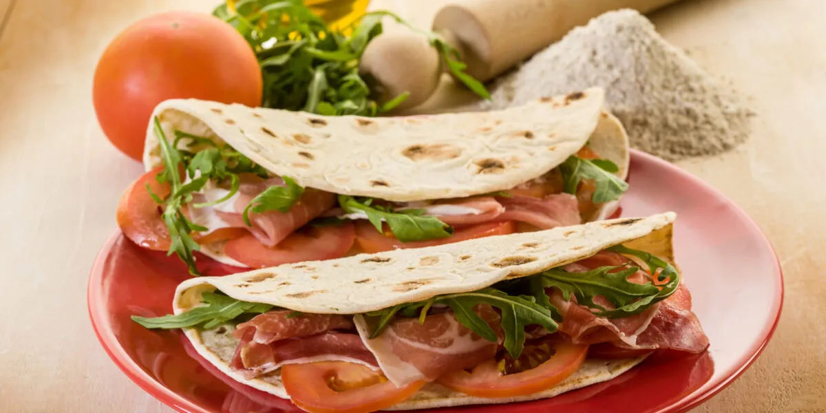 Piadine fit fatte in casa farcite con prosciutto crudo, pomodoro e rucola, servite su un piatto rosso.