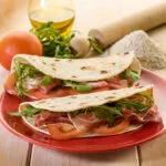 Piadine fit fatte in casa farcite con prosciutto crudo, pomodoro e rucola, servite su un piatto rosso.