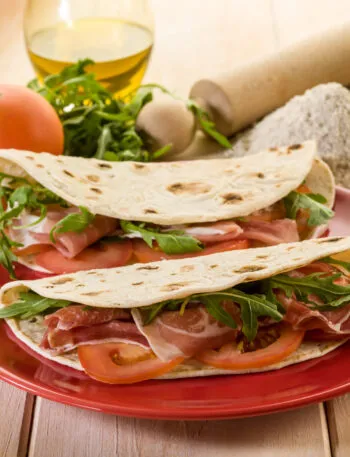 Piadine fit fatte in casa farcite con prosciutto crudo, pomodoro e rucola, servite su un piatto rosso.