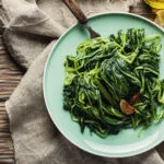 Spinaci saltati in padella con aglio e peperoncino serviti in un piatto verde su tavolo di legno.