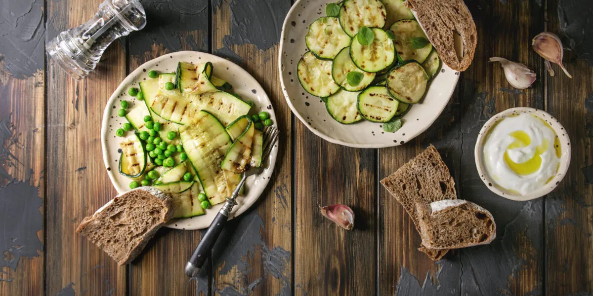 Piatti di zucchine grigliate con piselli, pane integrale e salsa allo yogurt su un tavolo rustico in legno.