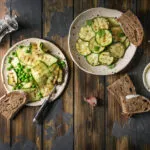 Piatti di zucchine grigliate con piselli, pane integrale e salsa allo yogurt su un tavolo rustico in legno.