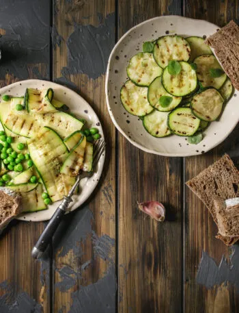 Piatti di zucchine grigliate con piselli, pane integrale e salsa allo yogurt su un tavolo rustico in legno.