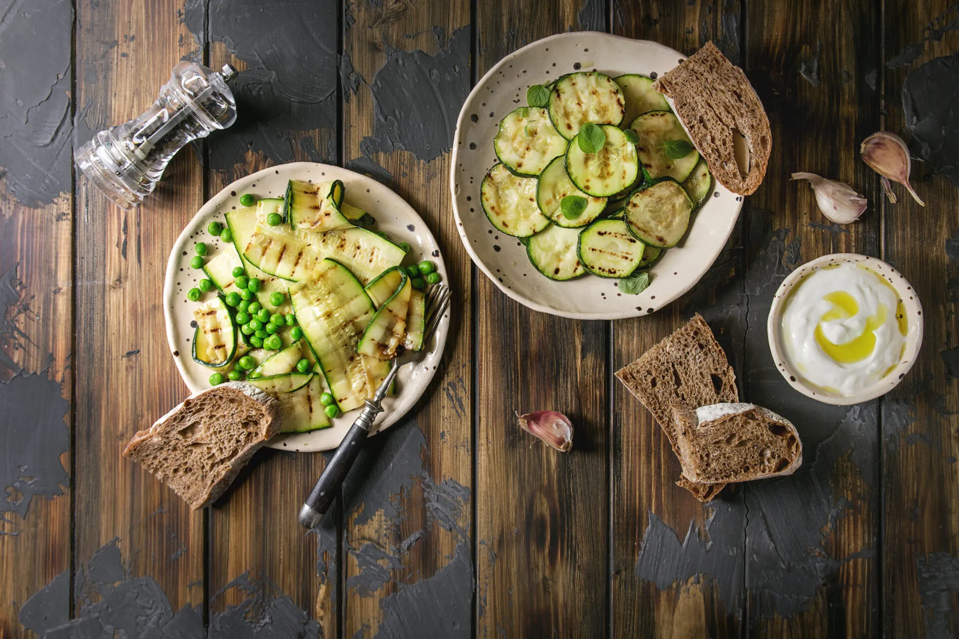 Piatti di zucchine grigliate con piselli, pane integrale e salsa allo yogurt su un tavolo rustico in legno.