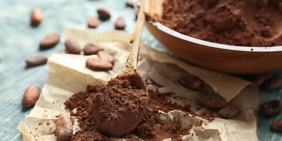 Cacao amaro in polvere su un cucchiaio di legno, con ciotola e fave di cacao su un tavolo azzurro.