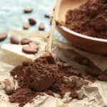 Cacao amaro in polvere su un cucchiaio di legno, con ciotola e fave di cacao su un tavolo azzurro.
